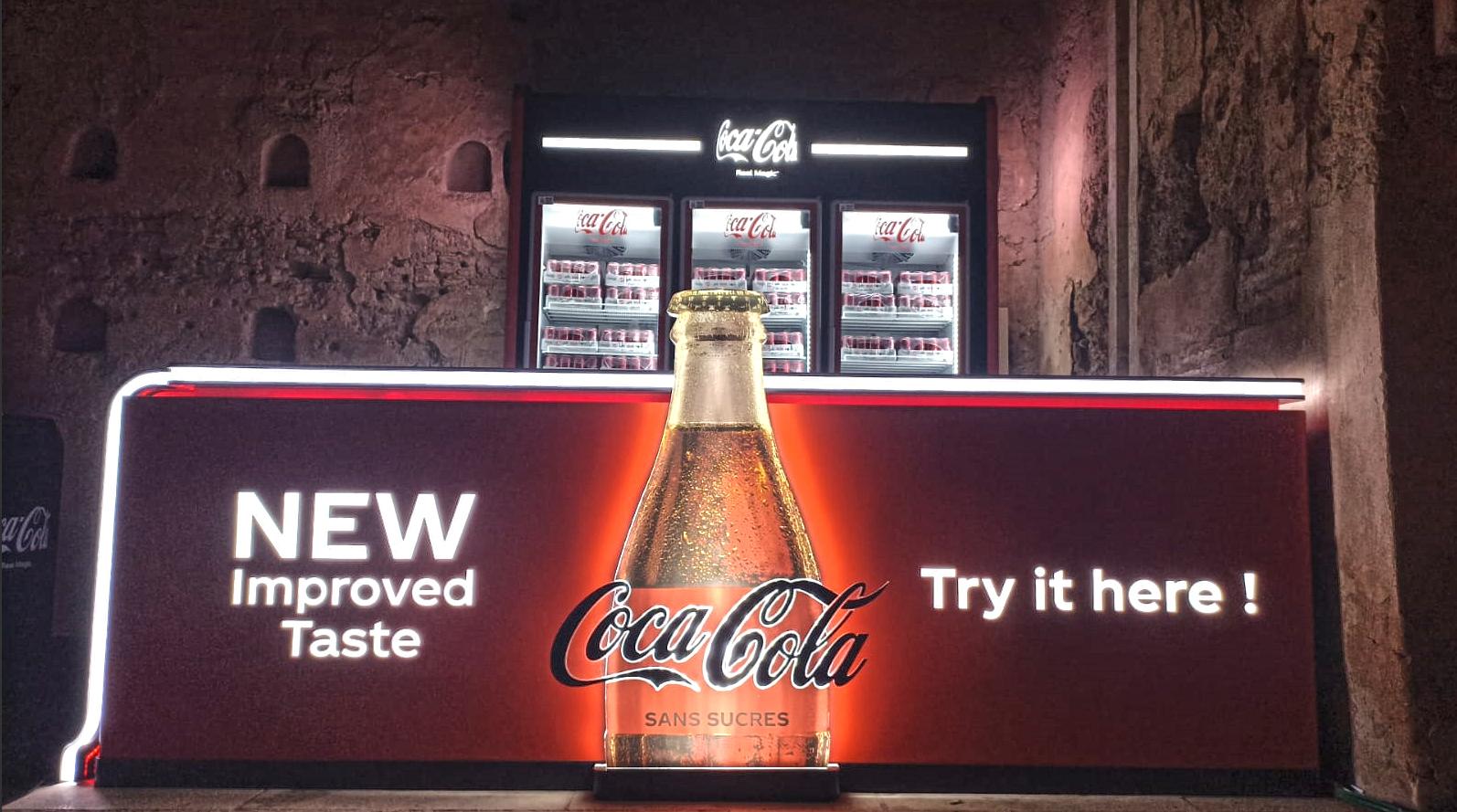 Comptoir-bar Coca-Cola sans sucres illuminé de nuit, bouteille rétroéclairée et réfrigérateurs vitrés, devant un mur en pisé.