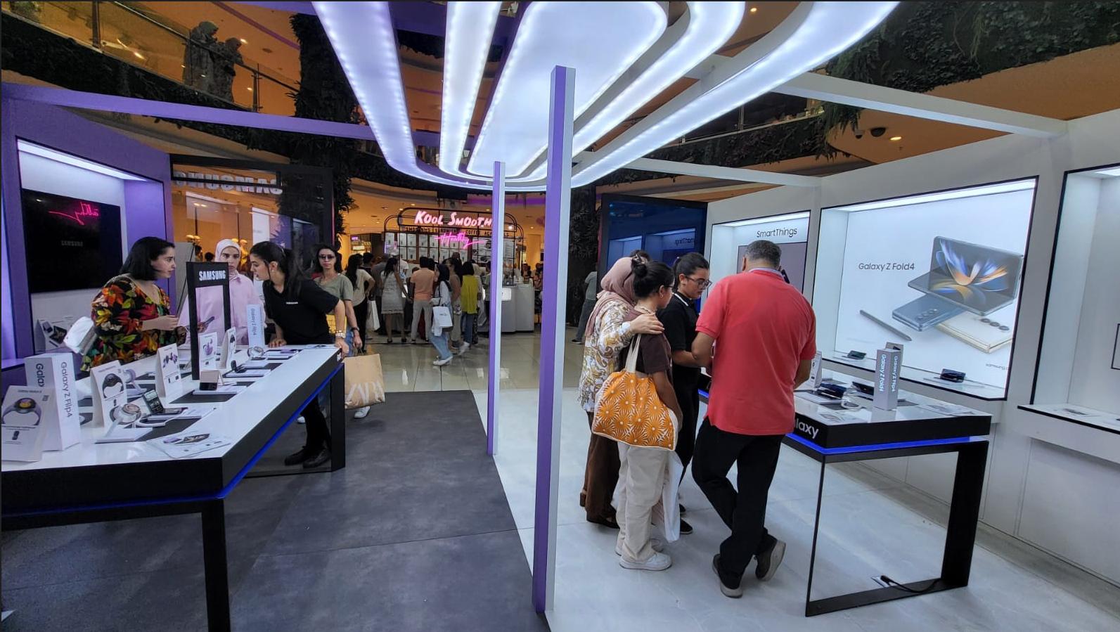 Visiteurs aux tables de démonstration du kiosque Samsung, sous un plafond lumineux ondulé.