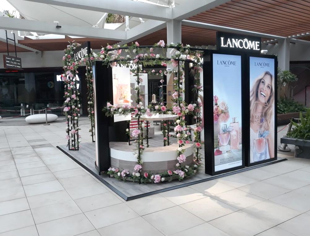 Pop-up Lancôme sous une pergola couverte de roses, avec comptoir arrondi et caissons lumineux.