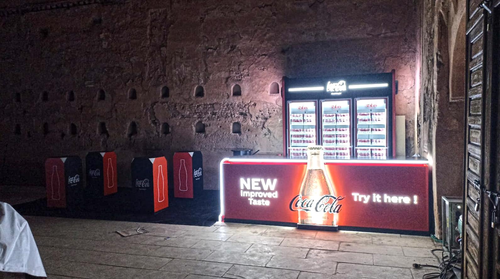 Vue large de nuit : le comptoir lumineux et les assises cubiques Coca-Cola alignées le long du mur.
